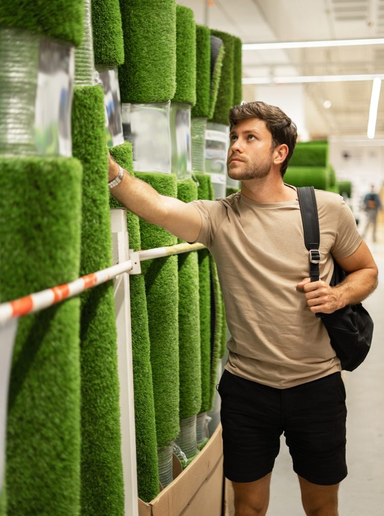 Casual man shopping in department of flooring and house goods while choosing rolled artificial grass